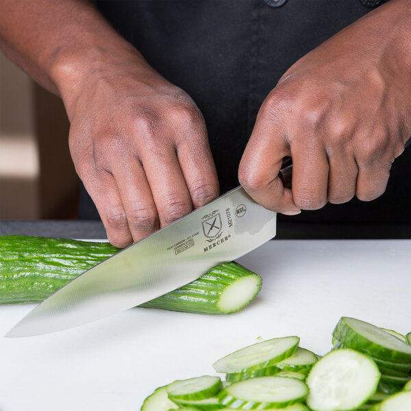 A person using a Mercer Culinary Genesis chef knife to cut a cucumber on a cutting board.