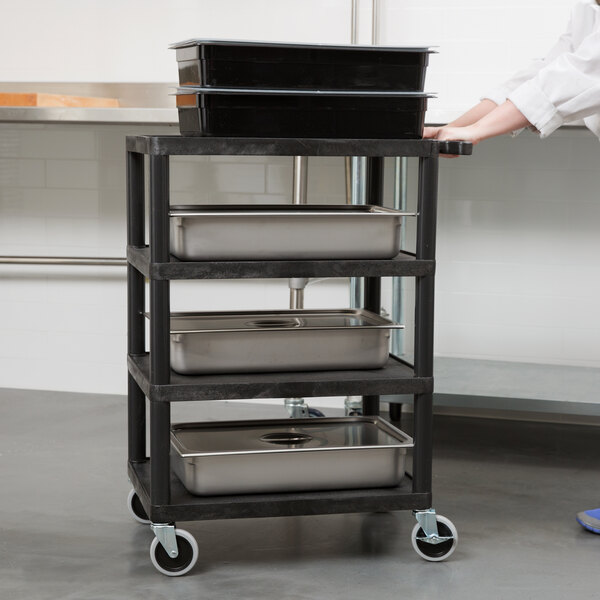 A person pushing a Luxor black metal serving cart with food containers on metal trays.