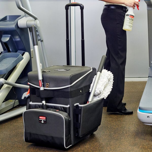 A woman using a black and grey Rubbermaid Executive Quick Cart to clean a gym room.