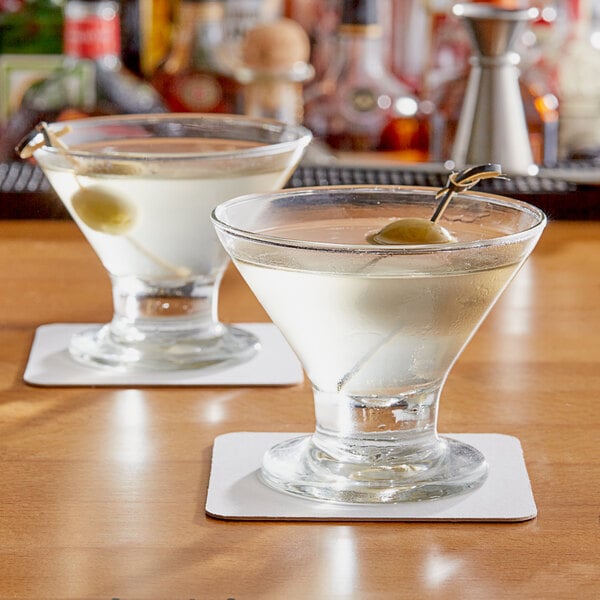 Two Acopa footed martini glasses with liquid and green olives on a table.