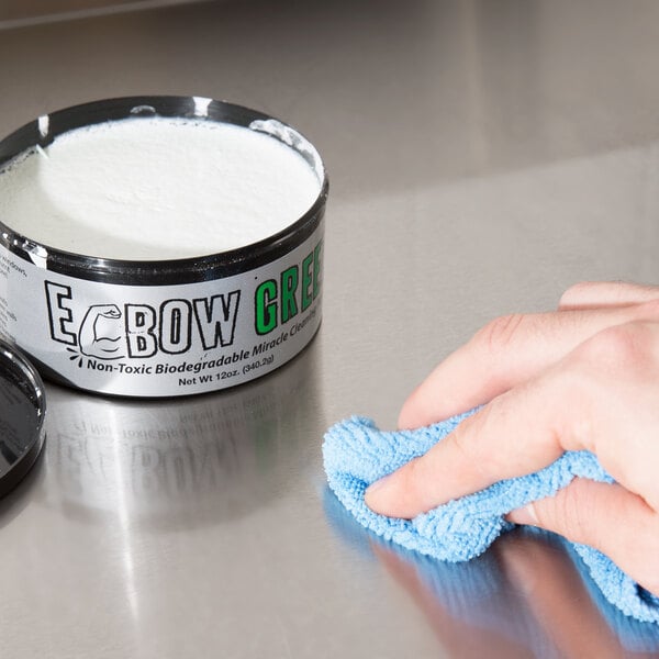 A hand wiping a professional kitchen counter with a blue cloth using white liquid from a can of Cres Cor Elbow Greez Miracle Cleaning Paste.