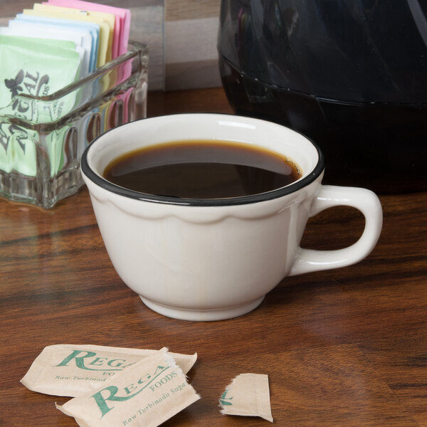 A CAC ivory china mug with a black band filled with coffee on a table with sugar packets.