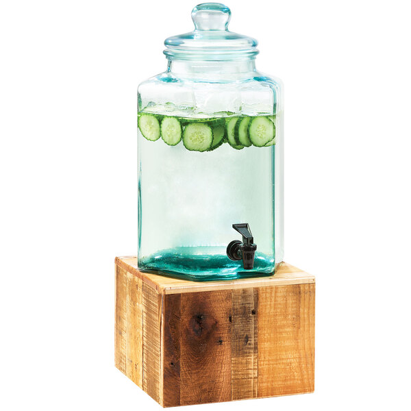 A Cal-Mil vintage glass beverage dispenser with cucumber water on a wooden stand.