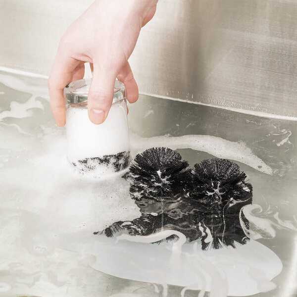 A hand using a Noble Products glass washer brush in a sink full of water to clean a glass.
