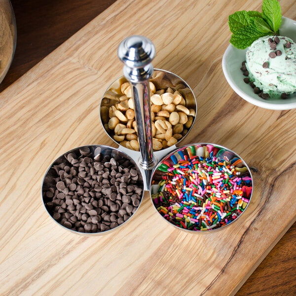 A Eastern Tabletop stainless steel condiment holder on a wood surface with bowls of sprinkles, chocolate chips, and nuts.