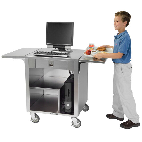 A young boy standing next to a Lakeside stainless steel cash register stand with a laptop on it.