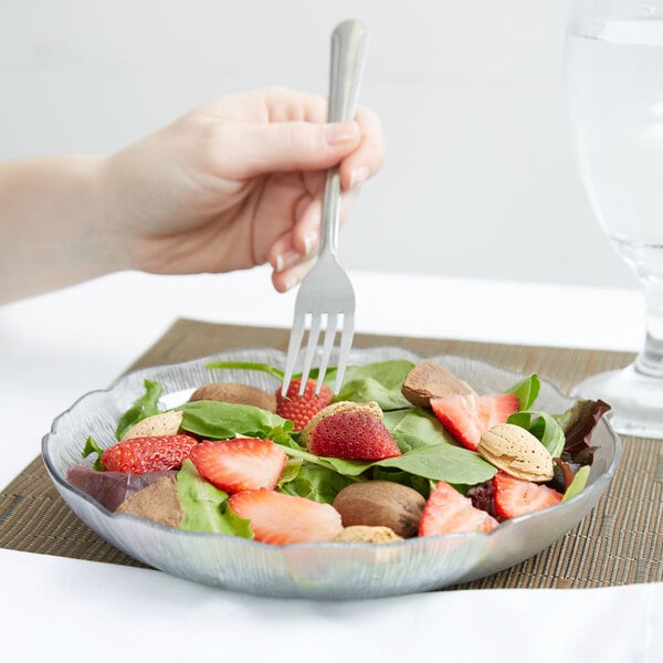 A hand holding a Carlisle clear polycarbonate plate with a fork over a bowl of salad.