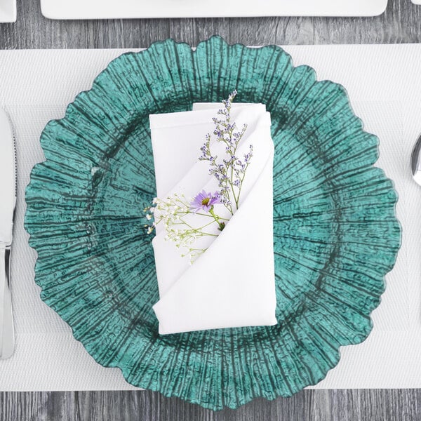 A teal Charge It by Jay glass charger plate with silverware and a white napkin with purple flowers on it.