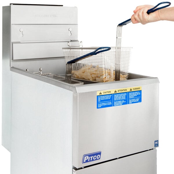 A hand holding a wire basket of food in front of a Pitco stainless steel floor fryer.