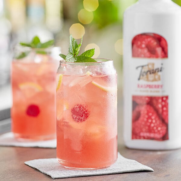 A glass of pink raspberry lemonade with mint leaves next to a white bottle of Torani Raspberry Puree Blend.