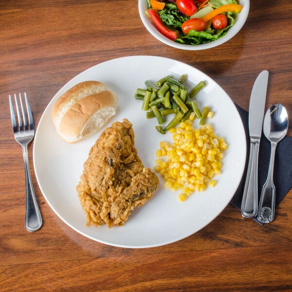 A CAC Majesty European bone china coupe plate with chicken, corn, and green beans on a table with a bowl of salad and a fork.
