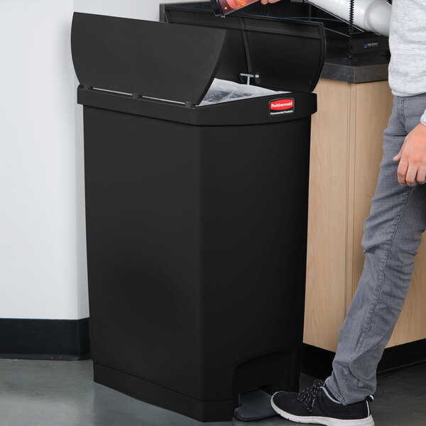 A man standing next to a black Rubbermaid Slim Jim end step-on trash can with the lid open.