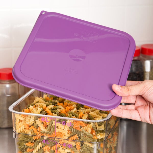 A person's hand holding a purple Cambro lid over a container of pasta.
