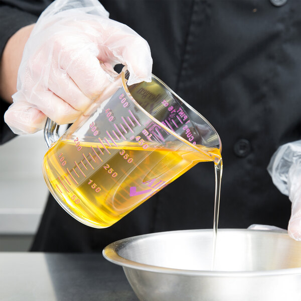 A person in a chef's uniform using a Cambro purple measuring cup to pour yellow liquid into a bowl.