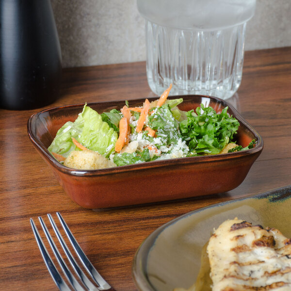 A Tuxton rectangular red rock china bowl with salad in it on a table next to a fork and a glass of water.