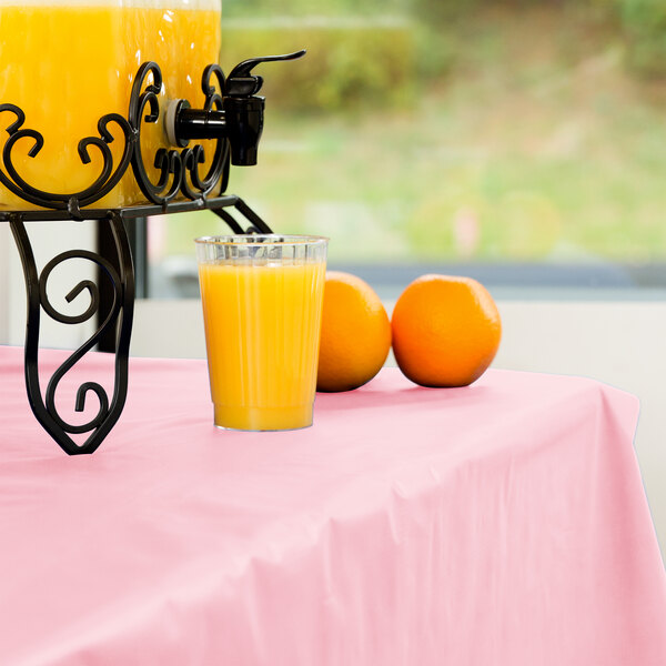 A glass of orange juice sits on a Classic Pink plastic table cover next to a drink dispenser with oranges.