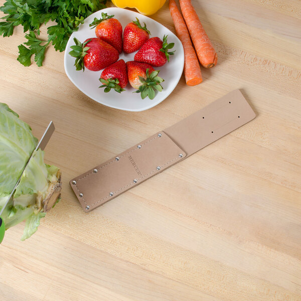 A Mercer Culinary Millennia leather sheath on a table with a Mercer Culinary produce knife, strawberries, and vegetables.