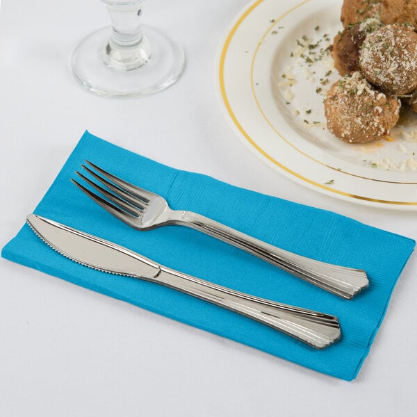 A fork and knife on a turquoise blue Creative Converting dinner napkin next to a plate of meatballs.