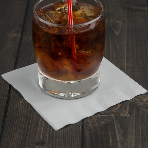 A glass of brown liquid with ice and a Creative Converting Shimmering Silver beverage napkin with a straw.