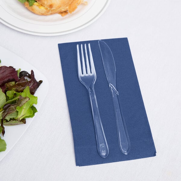 A fork and knife on a navy blue Creative Converting guest towel next to a plate of salad.