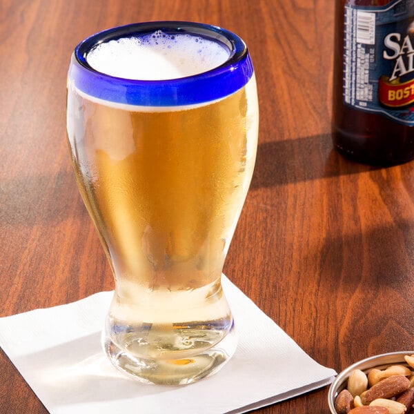 A Libbey Aruba pilsner glass with a cobalt blue rim filled with beer on a table with a bowl of mixed nuts.