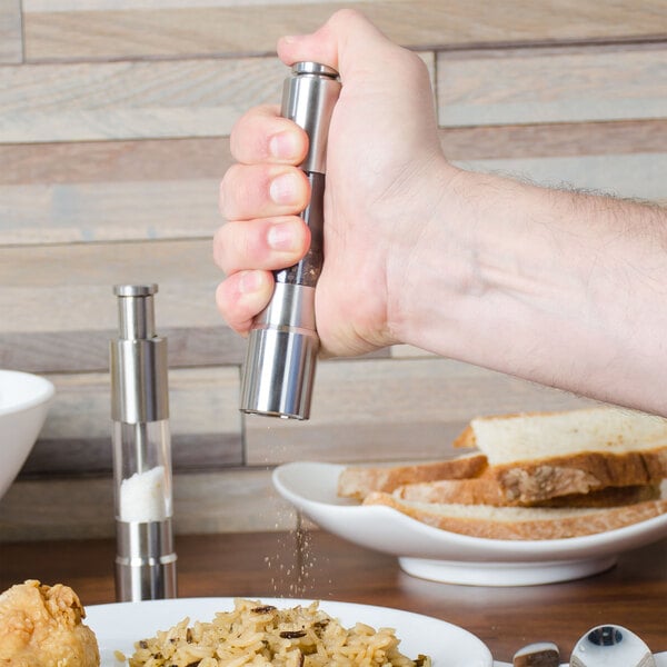 A hand holding an American Metalcraft stainless steel pepper mill over a plate of food.