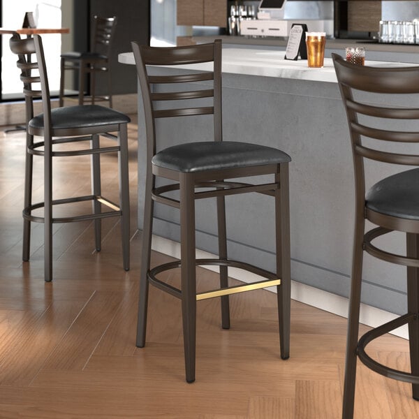 A group of Lancaster Table & Seating Spartan Series metal ladder back bar stools with black vinyl seats at a counter.