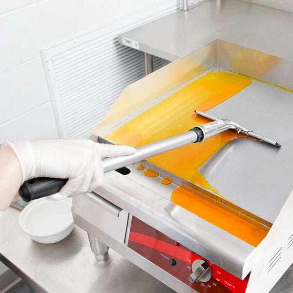 A gloved hand uses a 3M Scotch-Brite Griddle Squeegee on a metal surface.