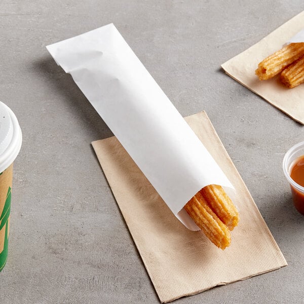 A churro in a white paper wrapper on a table with a cup of sauce and napkins.