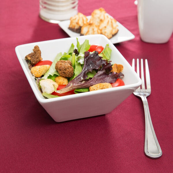 A bowl of salad with croutons and tomatoes in a white Libbey square porcelain bowl with a fork and knife.