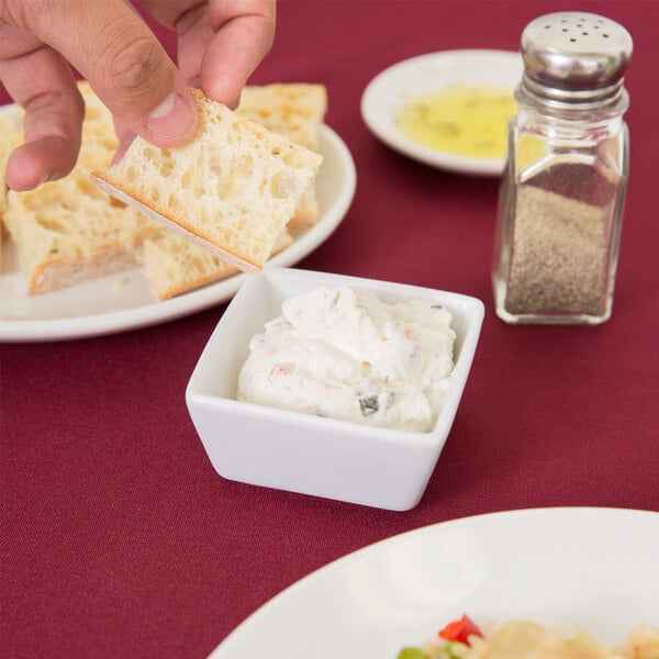 A person dipping a piece of bread into a white square bowl with dip.