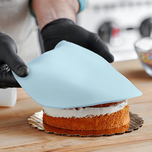 A person wearing black gloves uses a blue cloth to smooth blue Satin Ice fondant on a cake.