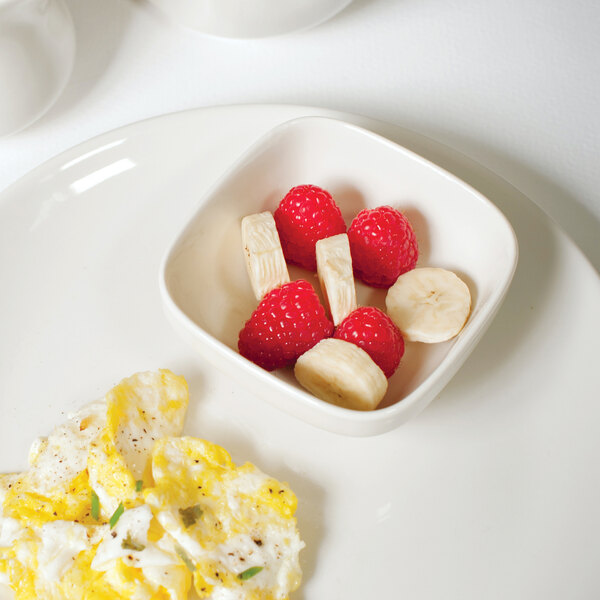 A Tuxton eggshell white square china bowl with raspberries, a banana, and an egg.