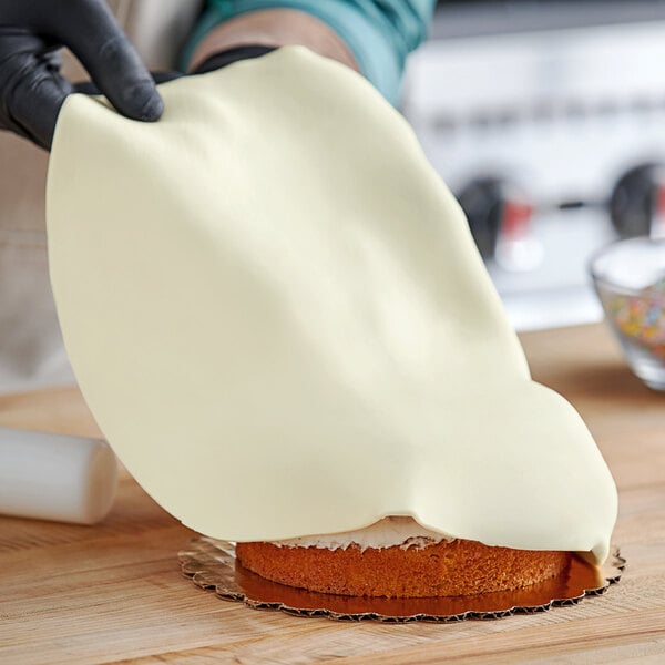 A person in gloves using a knife to cut ivory Satin Ice fondant on a cake.