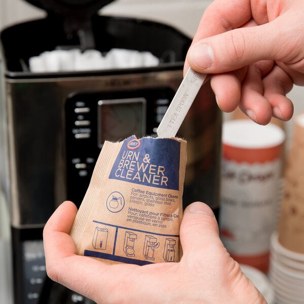 A hand holding a package of Urnex coffee urn cleaning powder.