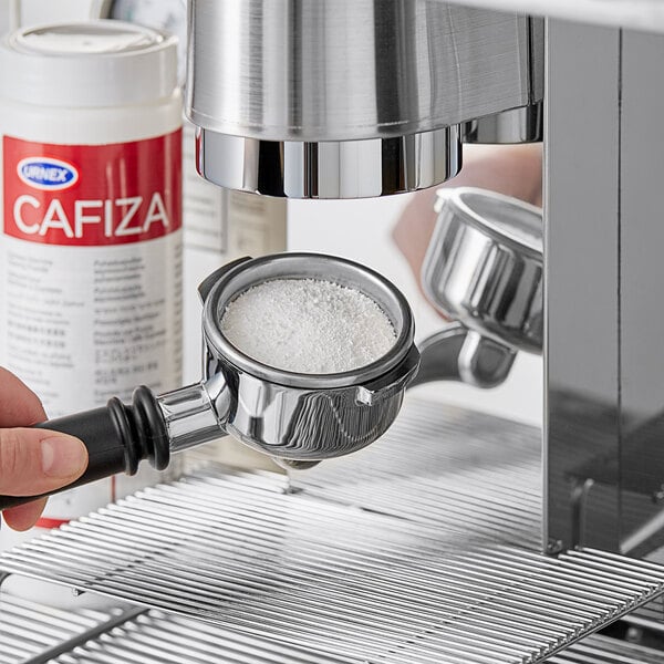 A hand pouring white powder from a silver container into a coffee machine.