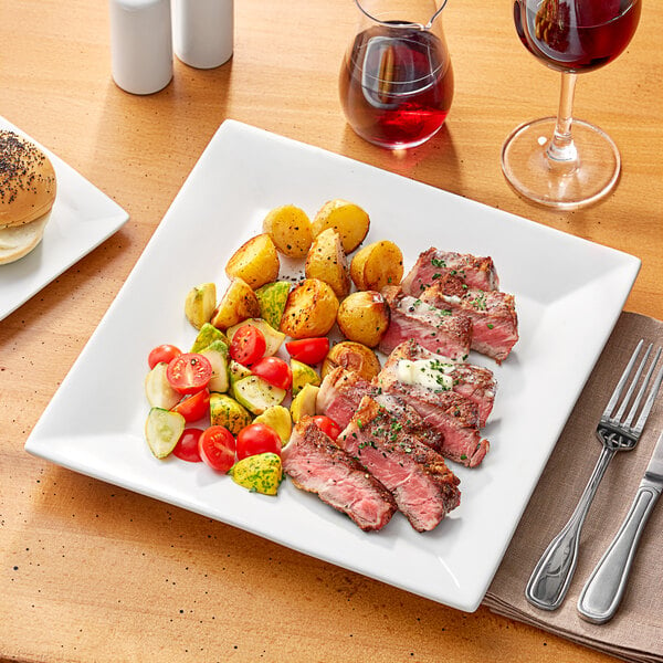 A plate of meat and vegetables with a fork and knife on a white Acopa square porcelain plate.