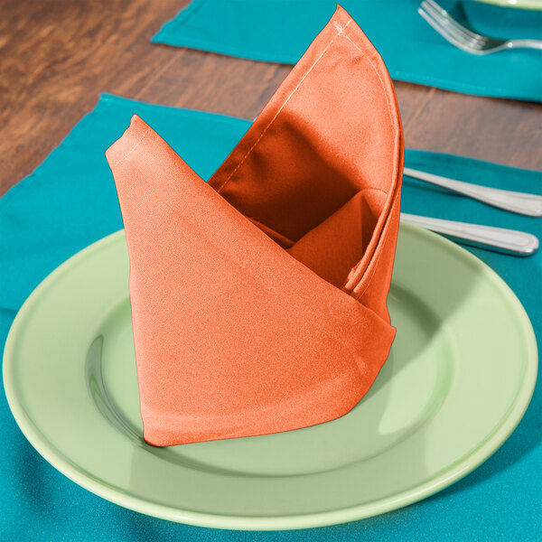 An orange Intedge cloth napkin folded on a plate with silverware.