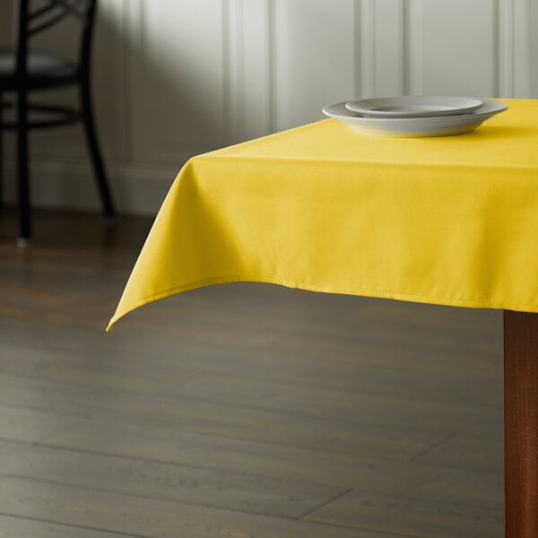 A square table with a yellow Intedge tablecloth on it and a plate.