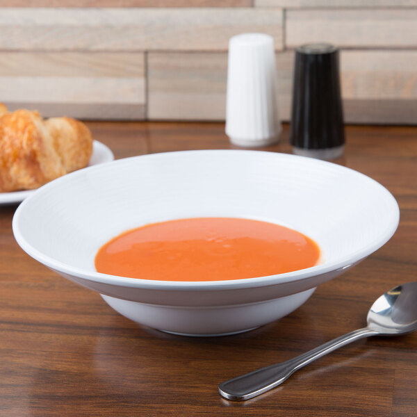 A white Minski melamine bowl of soup with a spoon on a table.