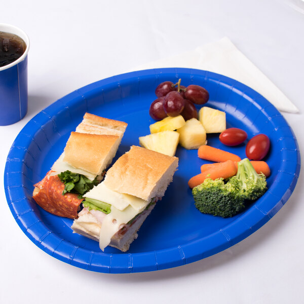 A Creative Converting cobalt blue oval paper platter with a sandwich, broccoli, and carrots on a table with a drink.