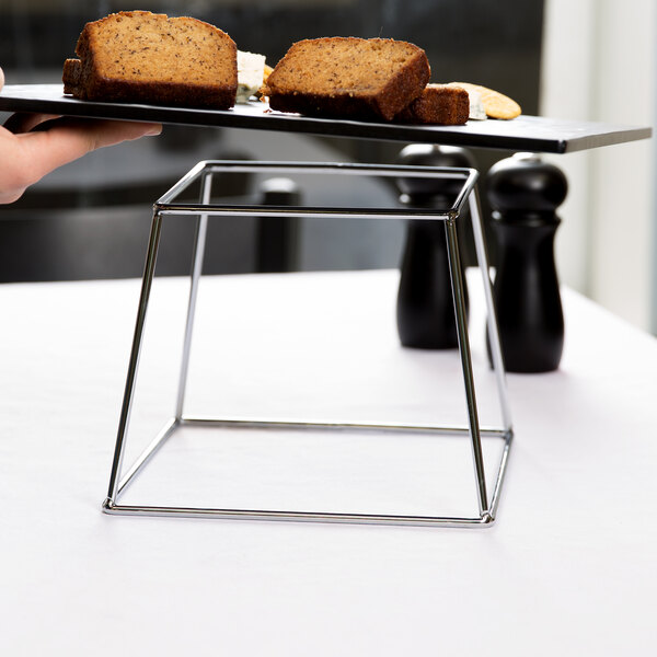 A hand holding a plate of bread on an American Metalcraft stainless steel square display stand.
