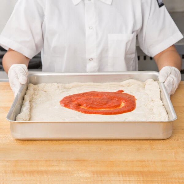 A person using an American Metalcraft aluminum pizza pan to hold pizza dough with red sauce.