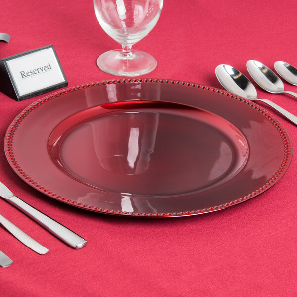 A red Charge It by Jay beaded plastic charger plate with silverware on a table.