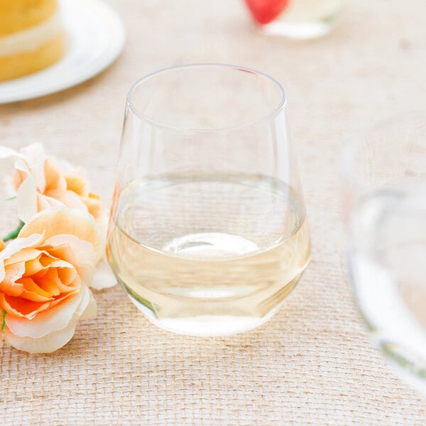 A clear plastic stemless wine goblet filled with white wine on a table with a cake.