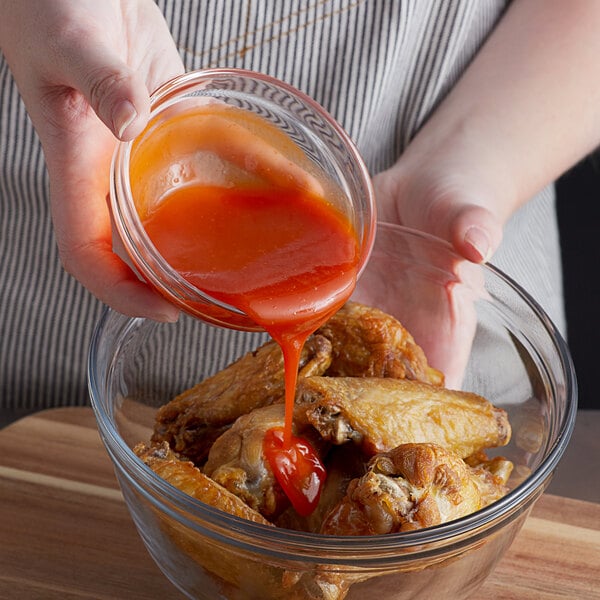 A hand pouring Crystal Chef's Recipe Garlic Hot Sauce into a bowl of chicken wings.
