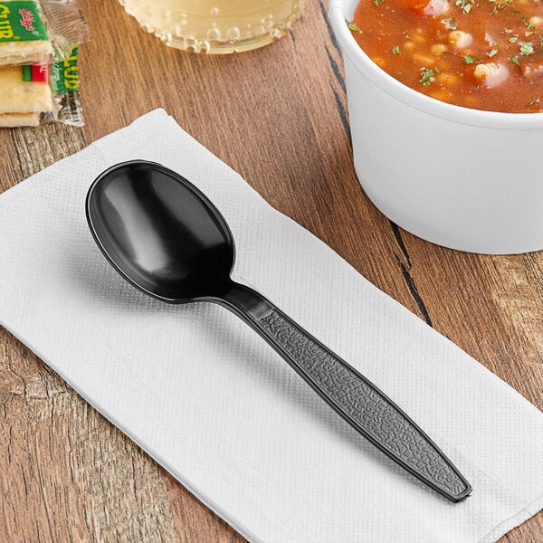 A black Visions heavy weight plastic soup spoon on a napkin next to a bowl of soup.