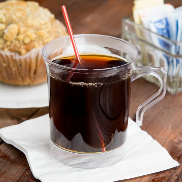 A Visions clear plastic coffee mug filled with coffee and a straw next to a muffin.