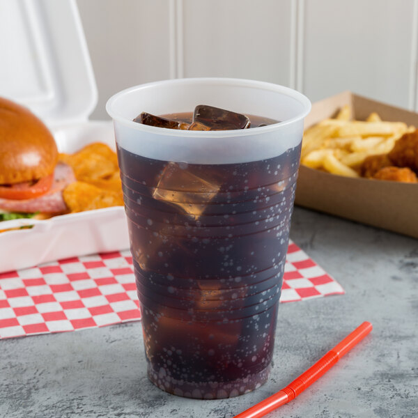 A Dart translucent plastic cup filled with soda and ice on a table with a sandwich and chicken.