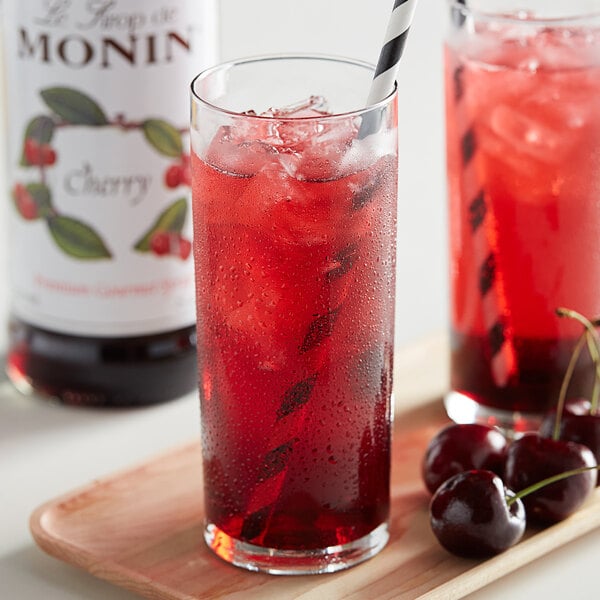 A glass of red cherry drink with ice and a straw with a bottle of Monin Premium Cherry Fruit Syrup.